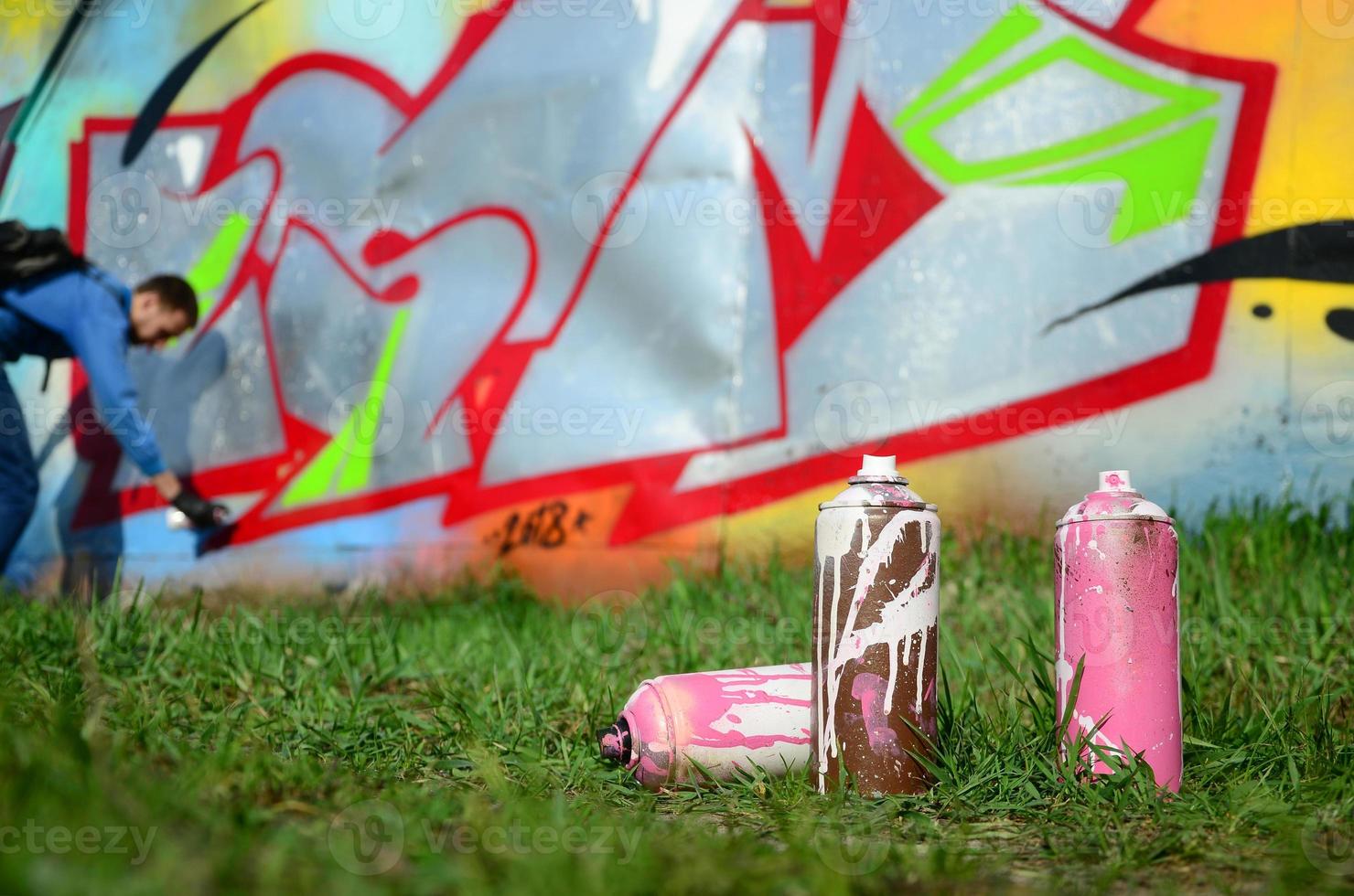 A few used paint cans against the background of the space with the wall on which the young guy draws a large graffiti drawing. Modern art of drawing walls in graffiti photo