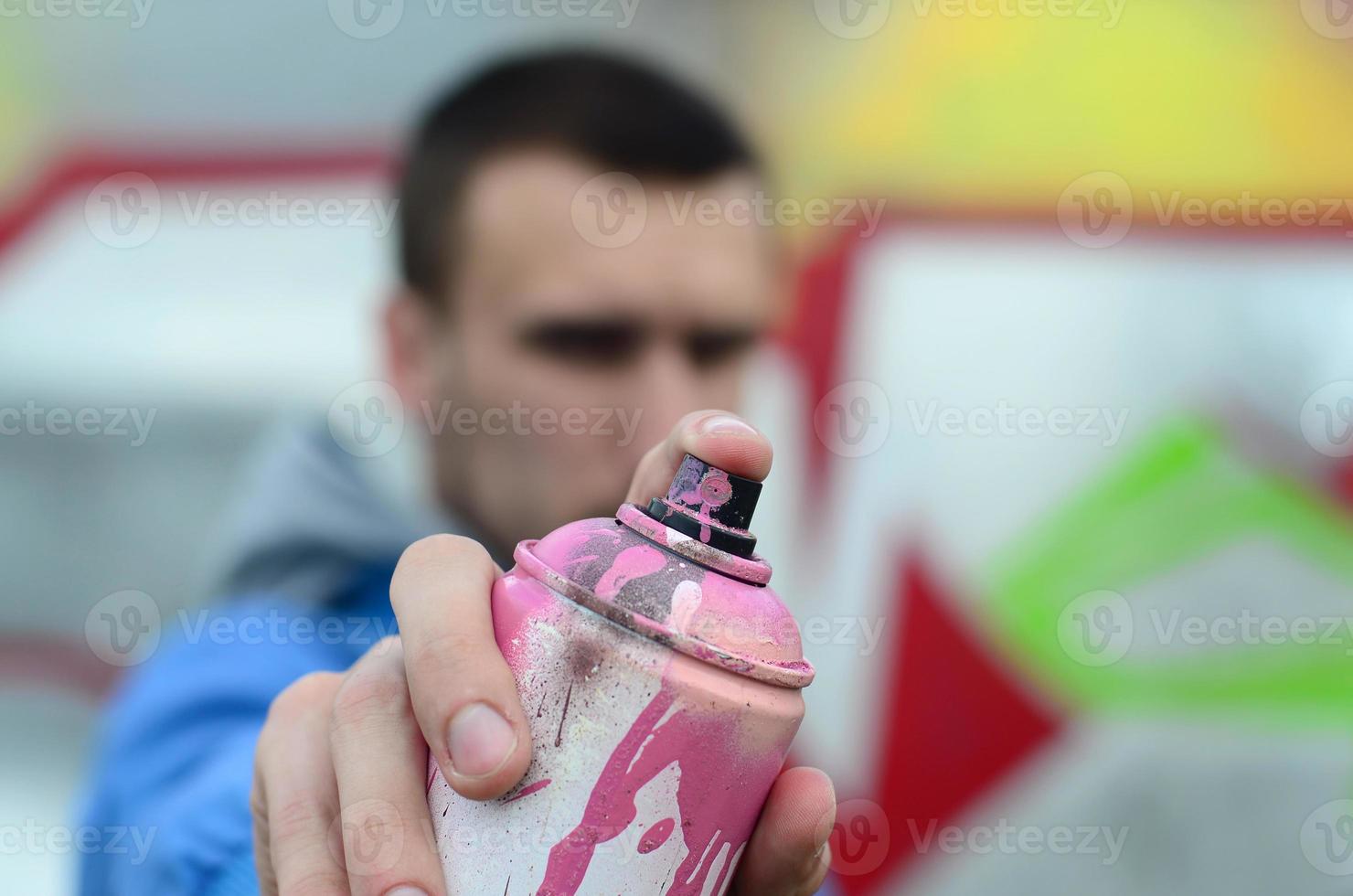 un joven artista de graffiti con una chaqueta azul sostiene una lata de pintura frente a él sobre un fondo de dibujo de graffiti de colores. concepto de arte callejero y vandalismo foto