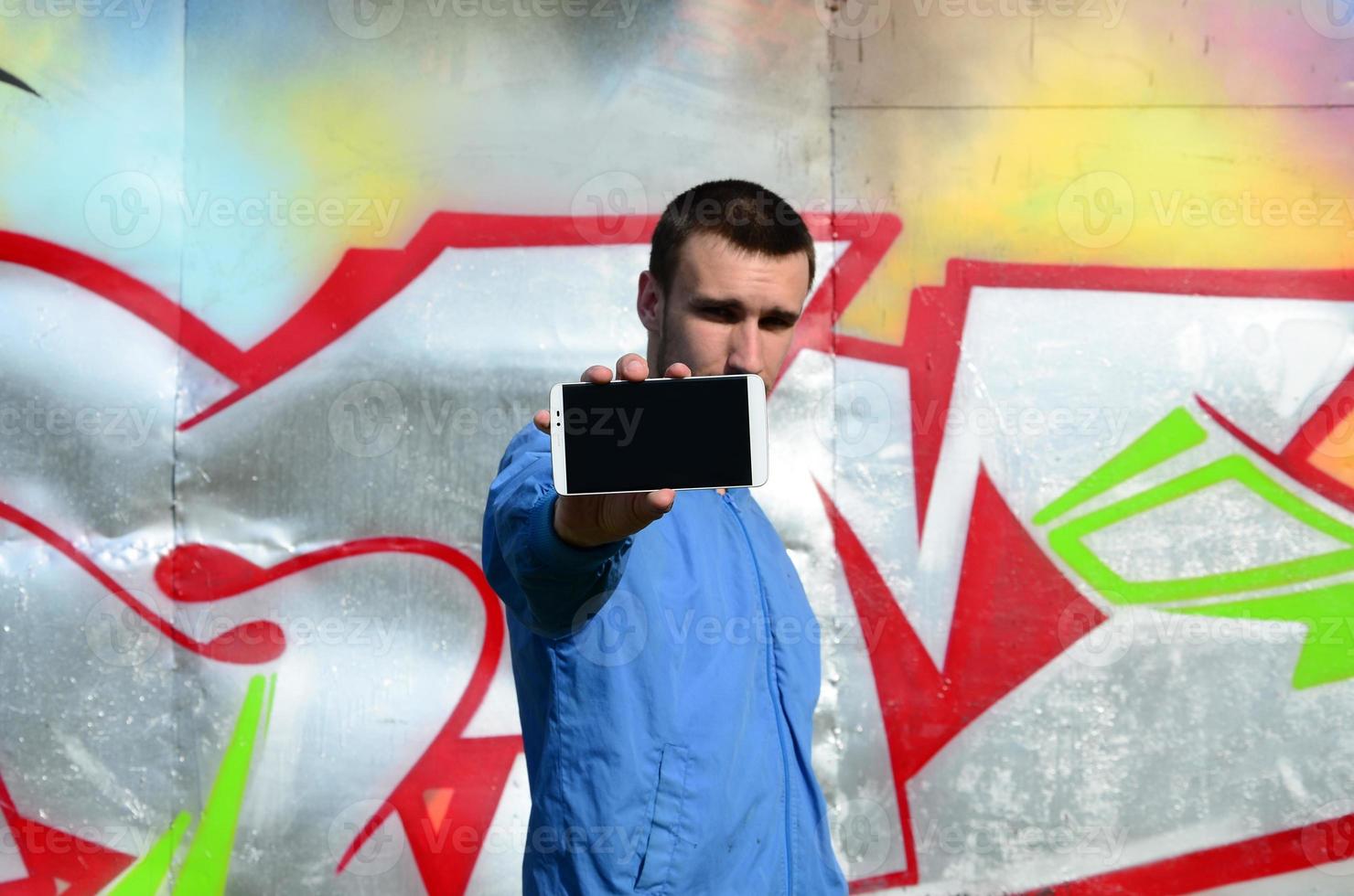 The graffiti artist demonstrates a smartphone with an empty black screen against the background of a colorful painted wall. Street art concept photo