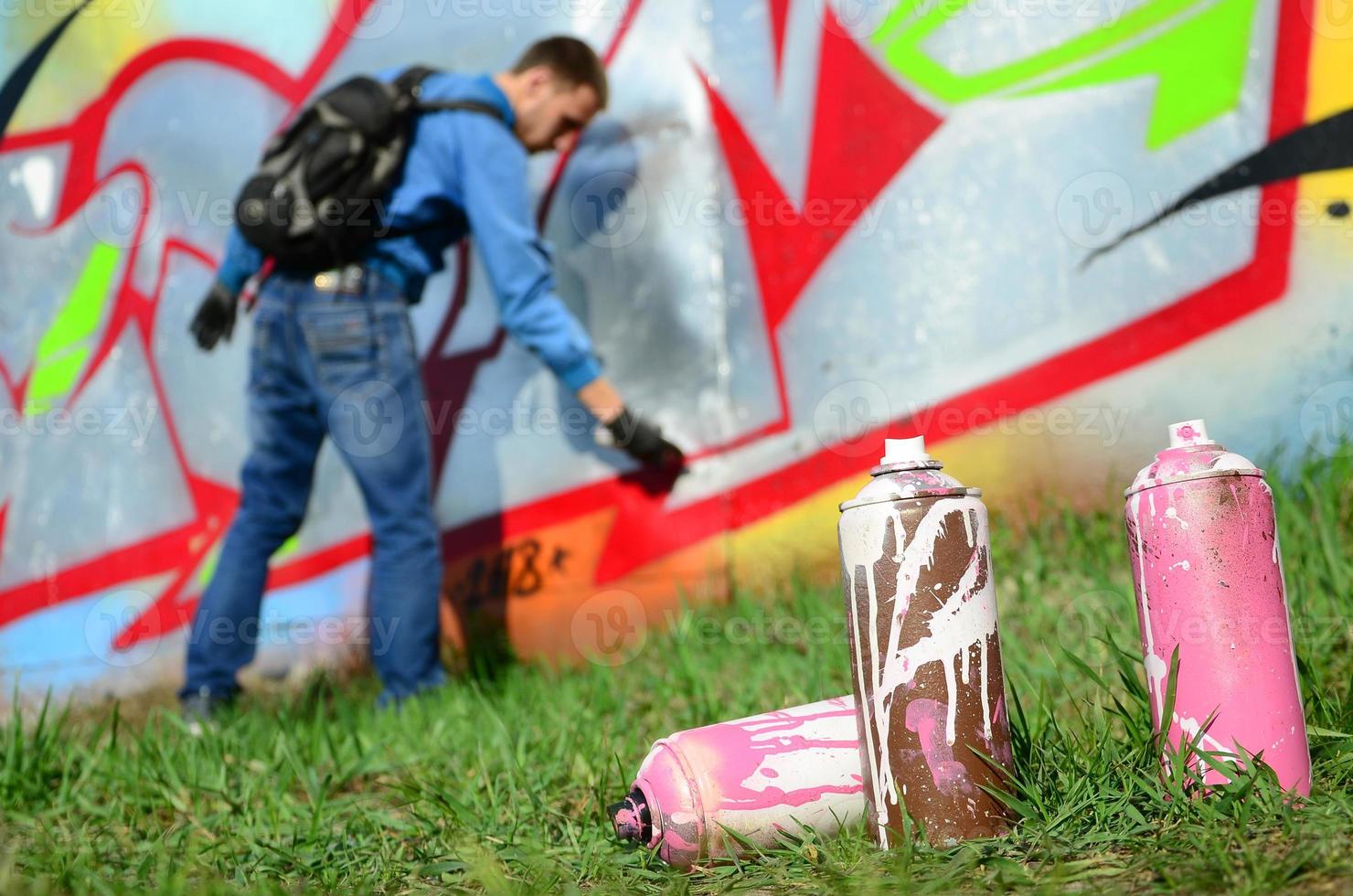 A few used paint cans against the background of the space with the wall on which the young guy draws a large graffiti drawing. Modern art of drawing walls in graffiti photo