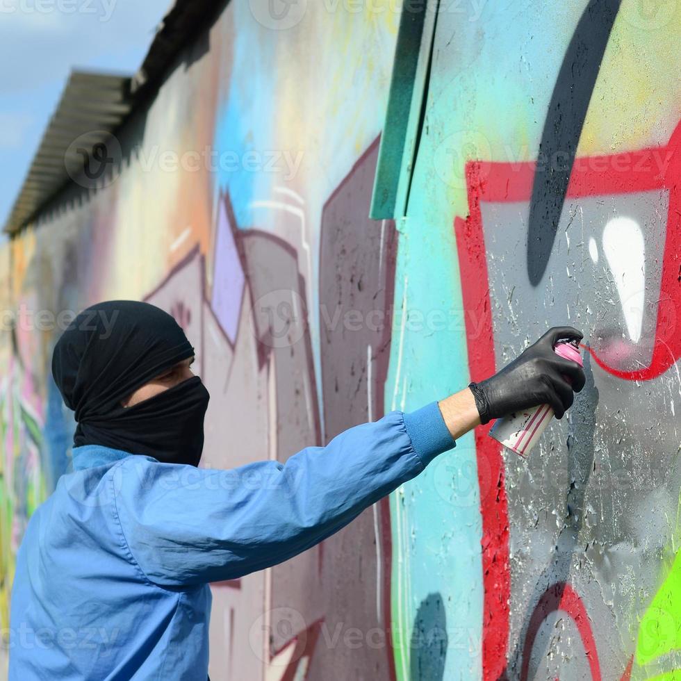 A young hooligan with a hidden face paints graffiti on a metal wall. Illegal vandalism concept photo