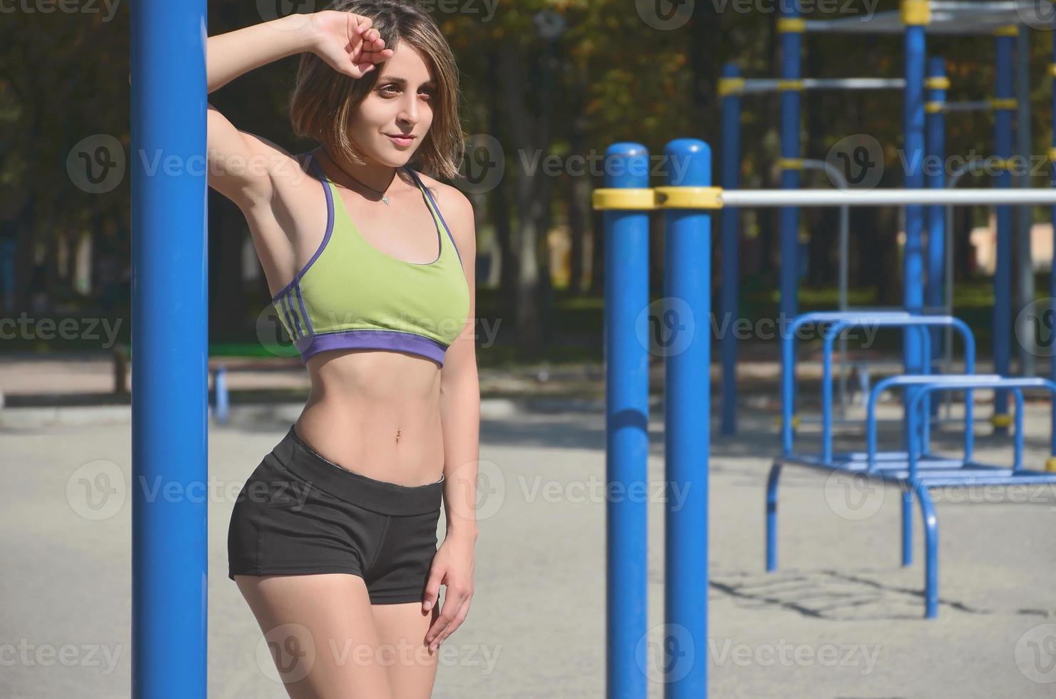 una joven atleta caucásica con una ropa deportiva verde brillante se alza contra el telón de fondo de un patio de recreo deportivo callejero para el entrenamiento de atletismo al aire libre. deportes de verano y concepto de estilo de vida saludable foto