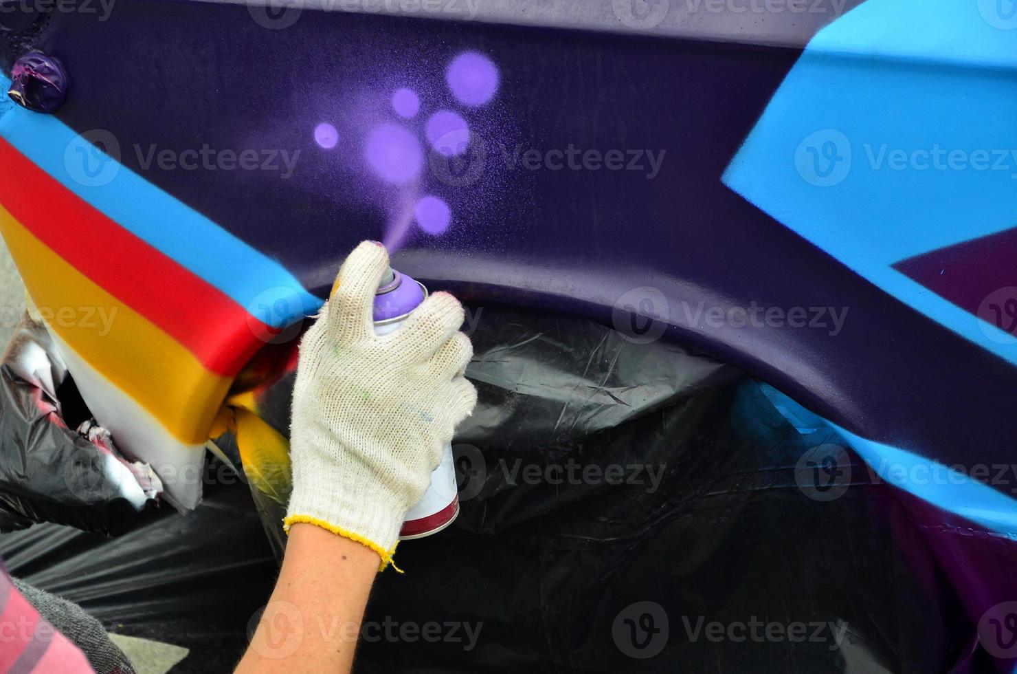 A hand with a spray can that draws a new colorful graffiti on the car. Photo of the process of drawing a graffiti on an automobile close-up. The concept of street art and illegal vandalism