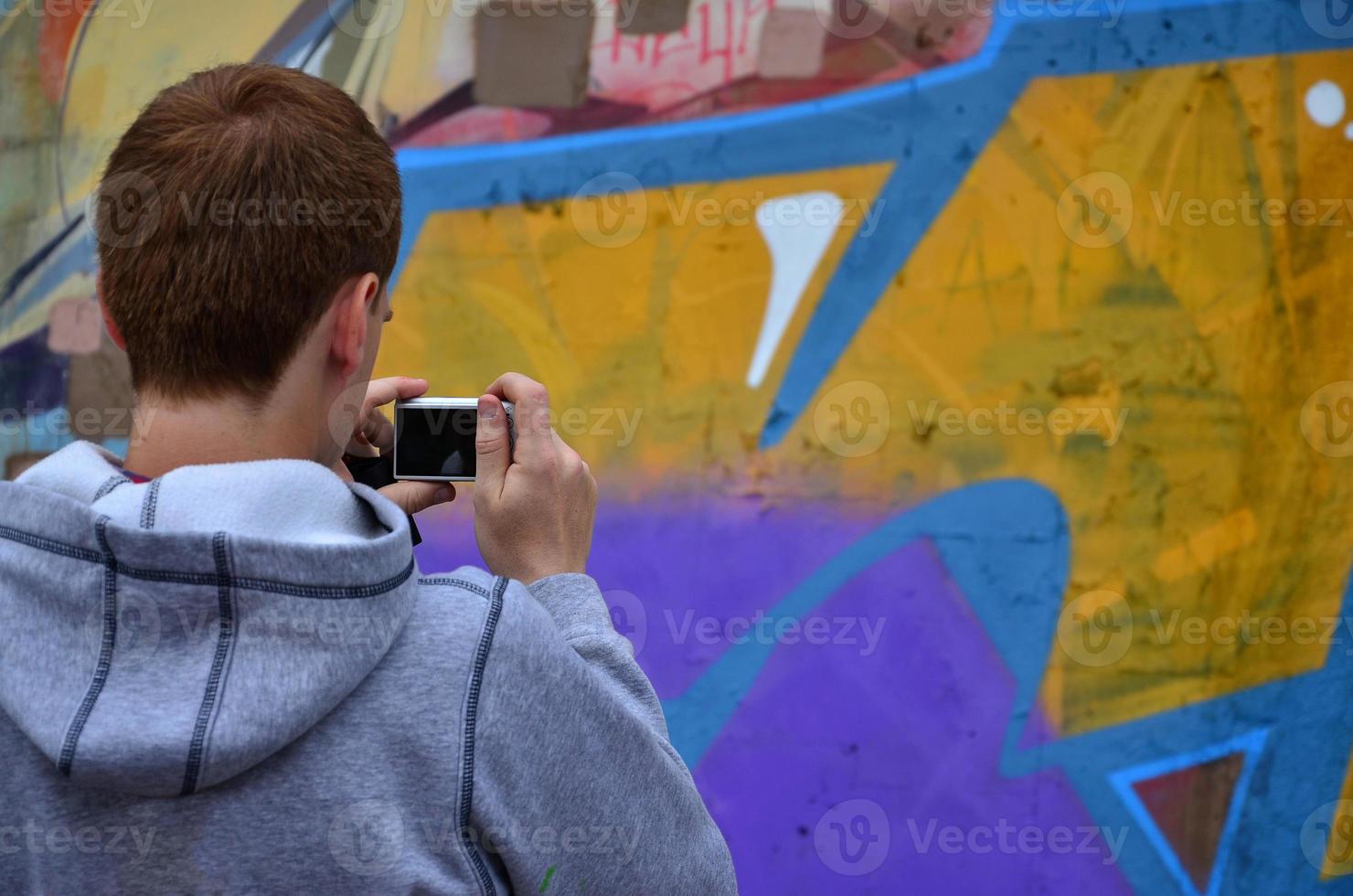 A young graffiti artist photographs his completed picture on the wall. The guy uses modern technology to capture a colorful abstract graffiti drawing. Focus on the photographing device photo