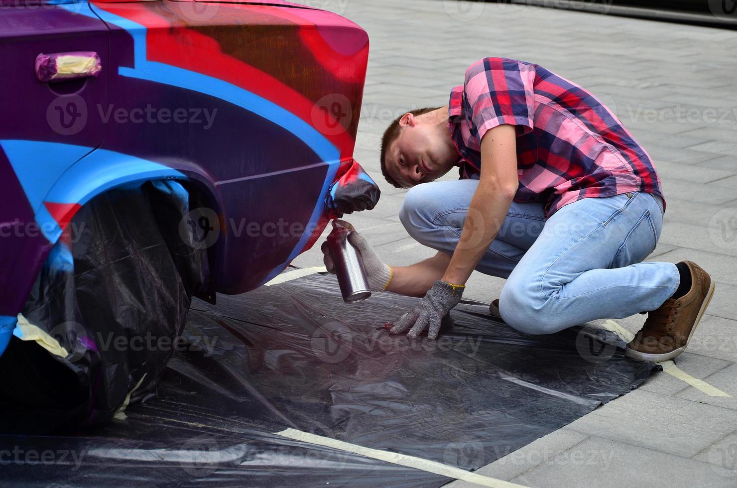 A young red-haired graffiti artist paints a new colorful graffiti on the car. Photo of the process of drawing a graffiti on a car close-up. The concept of street art and illegal vandalism
