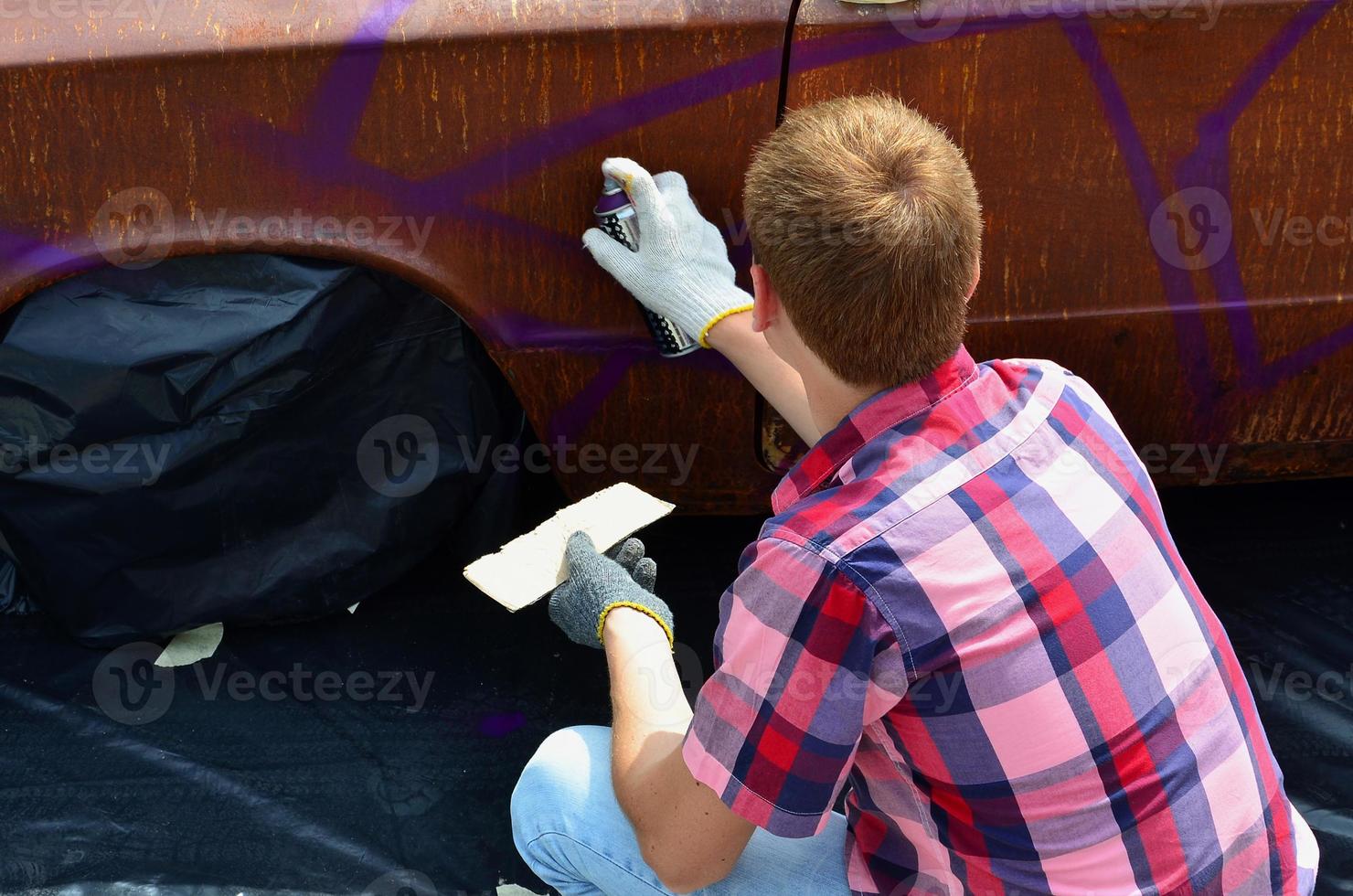 A young red-haired graffiti artist paints a new colorful graffiti on the car. Photo of the process of drawing a graffiti on a car close-up. The concept of street art and illegal vandalism