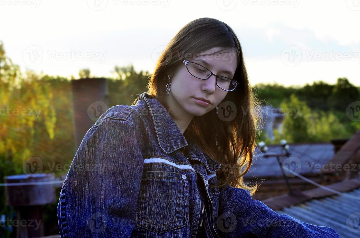 Girl in glasses and jeans jacket is resting on the roof of a hou photo