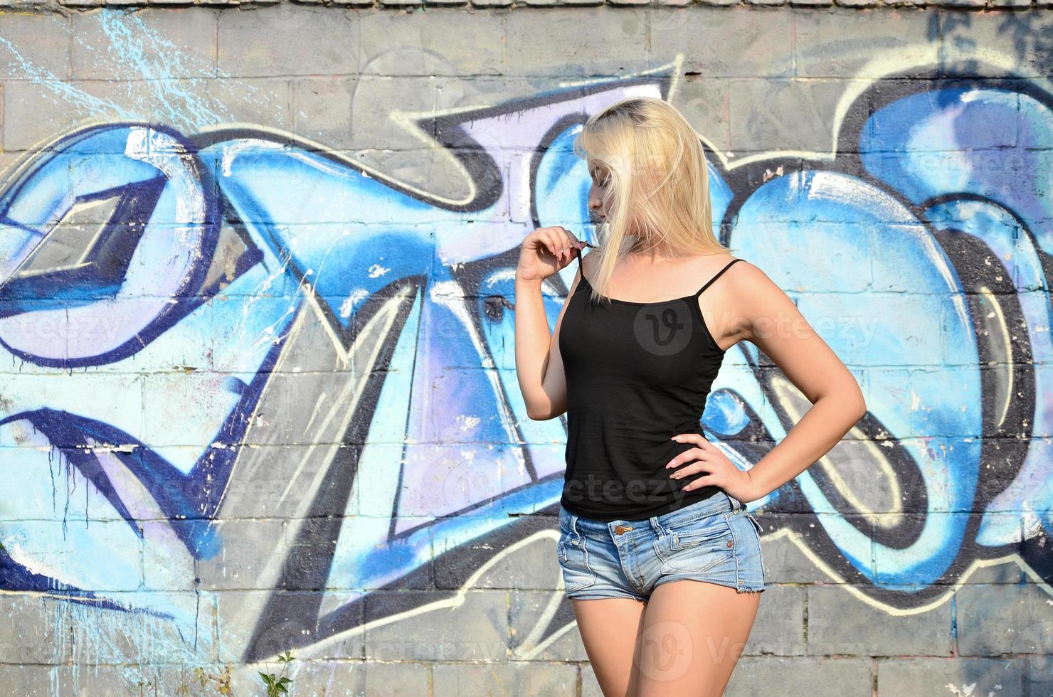 Sexy caucasian blonde girl in denim shorts and black tank top posing against graffiti wall in the daytime outdoors photo