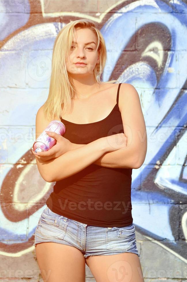 A young and beautiful sexy girl graffiti artist with a paint spray stands on the wall background with a graffiti pattern in blue and purple tones photo