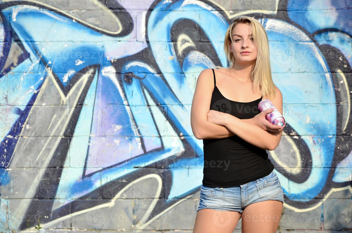 A young and beautiful sexy girl graffiti artist with a paint spray stands on the wall background with a graffiti pattern in blue and purple tones photo