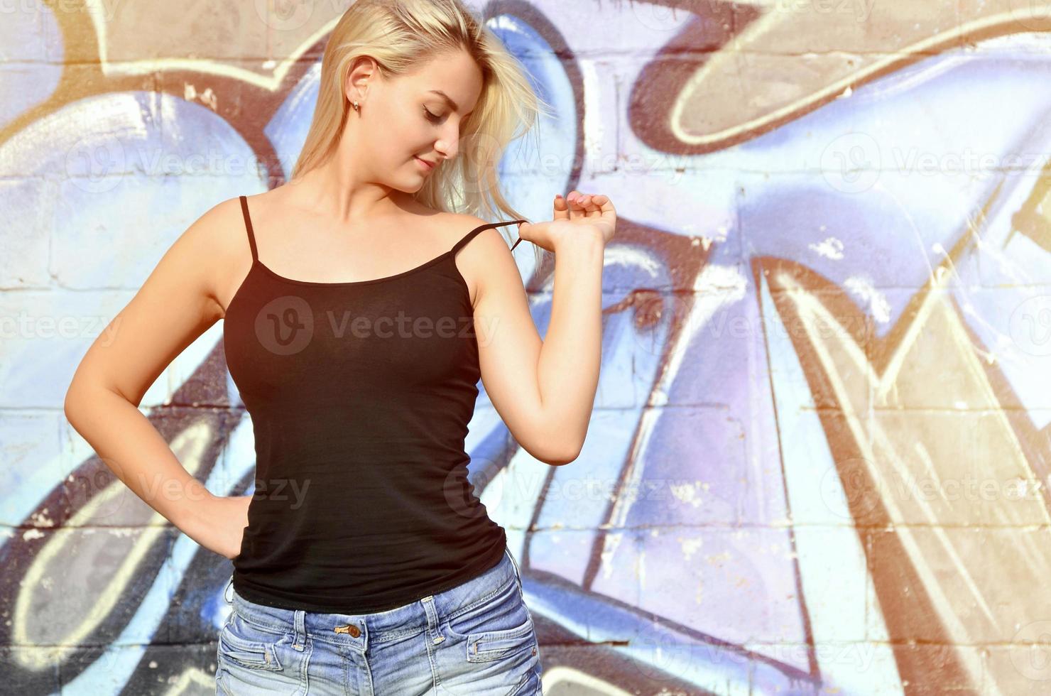 Chica rubia caucásica sexy en pantalones cortos de mezclilla y camiseta sin mangas negra posando contra la pared de graffiti durante el día al aire libre foto