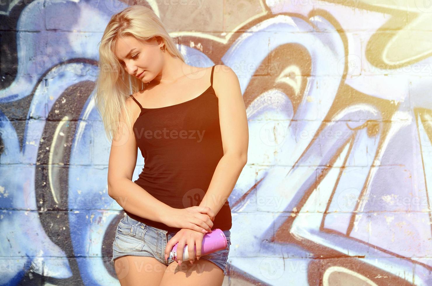 A young and beautiful sexy girl graffiti artist with a paint spray stands on the wall background with a graffiti pattern in blue and purple tones photo