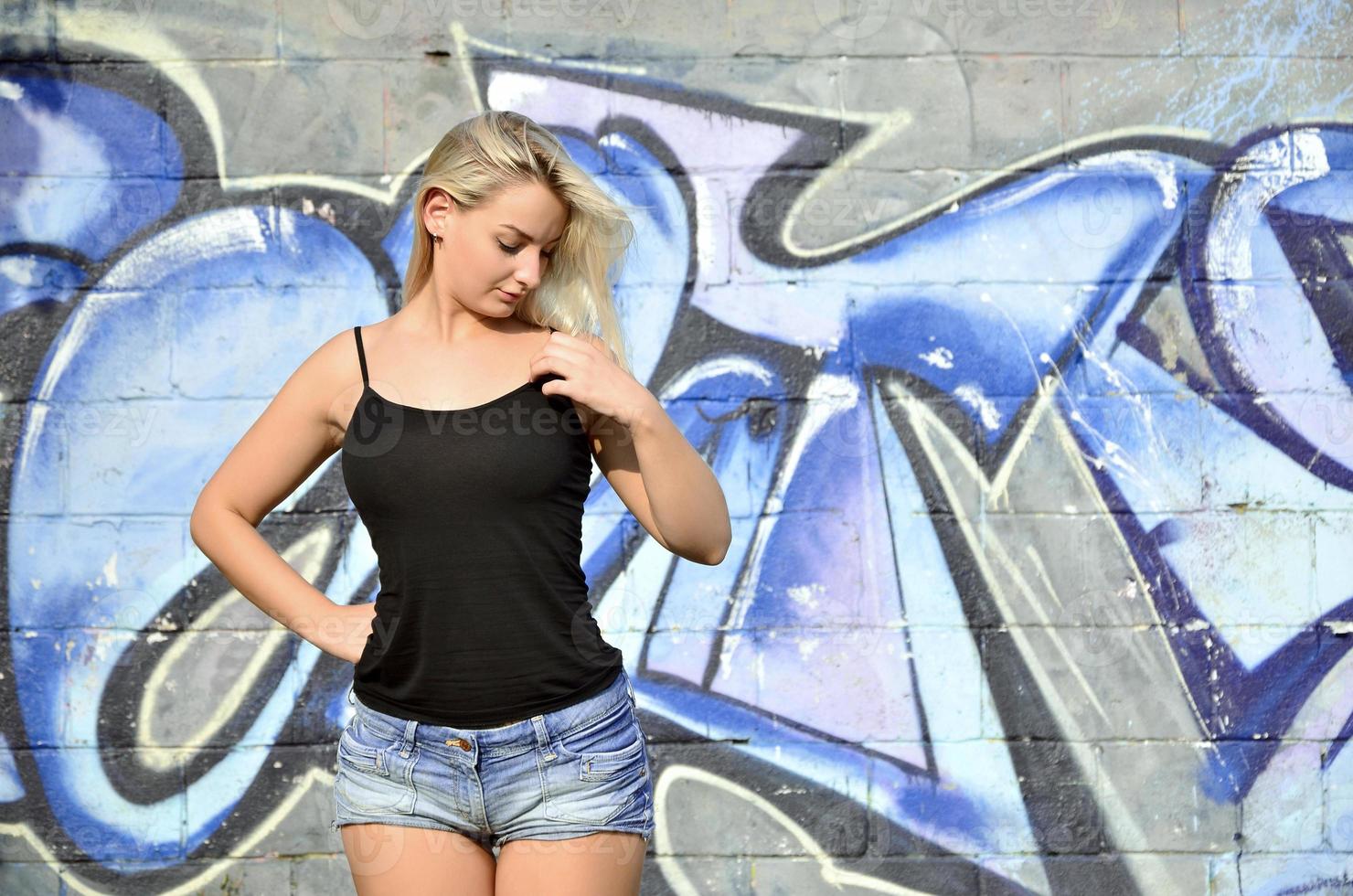 Chica rubia caucásica sexy en pantalones cortos de mezclilla y camiseta sin mangas negra posando contra la pared de graffiti durante el día al aire libre foto