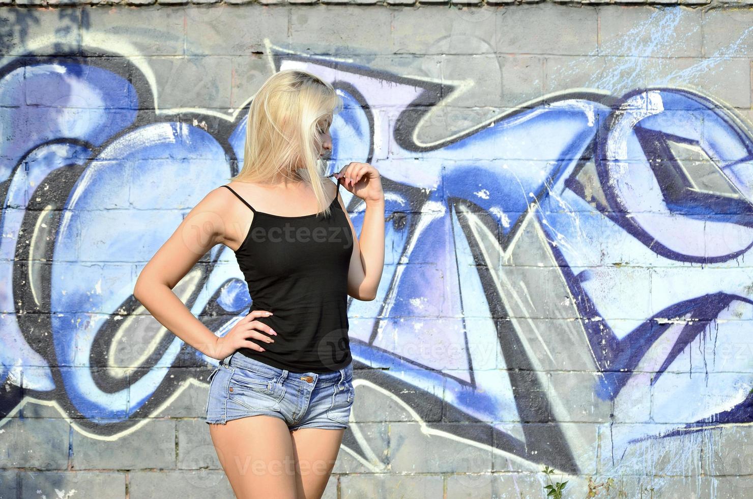 Sexy caucasian blonde girl in denim shorts and black tank top posing against graffiti wall in the daytime outdoors photo