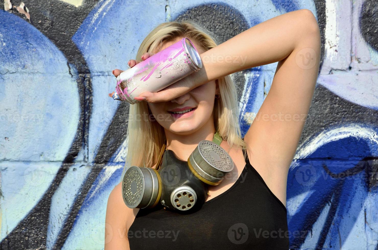 Young and beautiful smiling sexy girl graffiti artist with gas mask on her neck hiding his eyes with a spray can standing on a wall background with a graffiti pattern in blue and purple tones photo