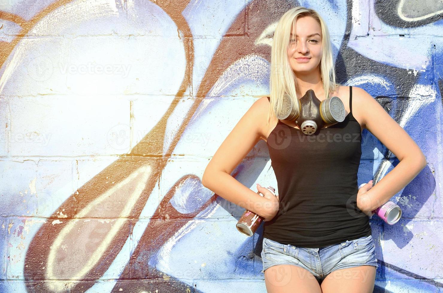 A smiling young and beautiful sexy girl graffiti artist with a paint spray and gas mask on her neck stands on the wall background with a graffiti pattern in blue and purple tones photo