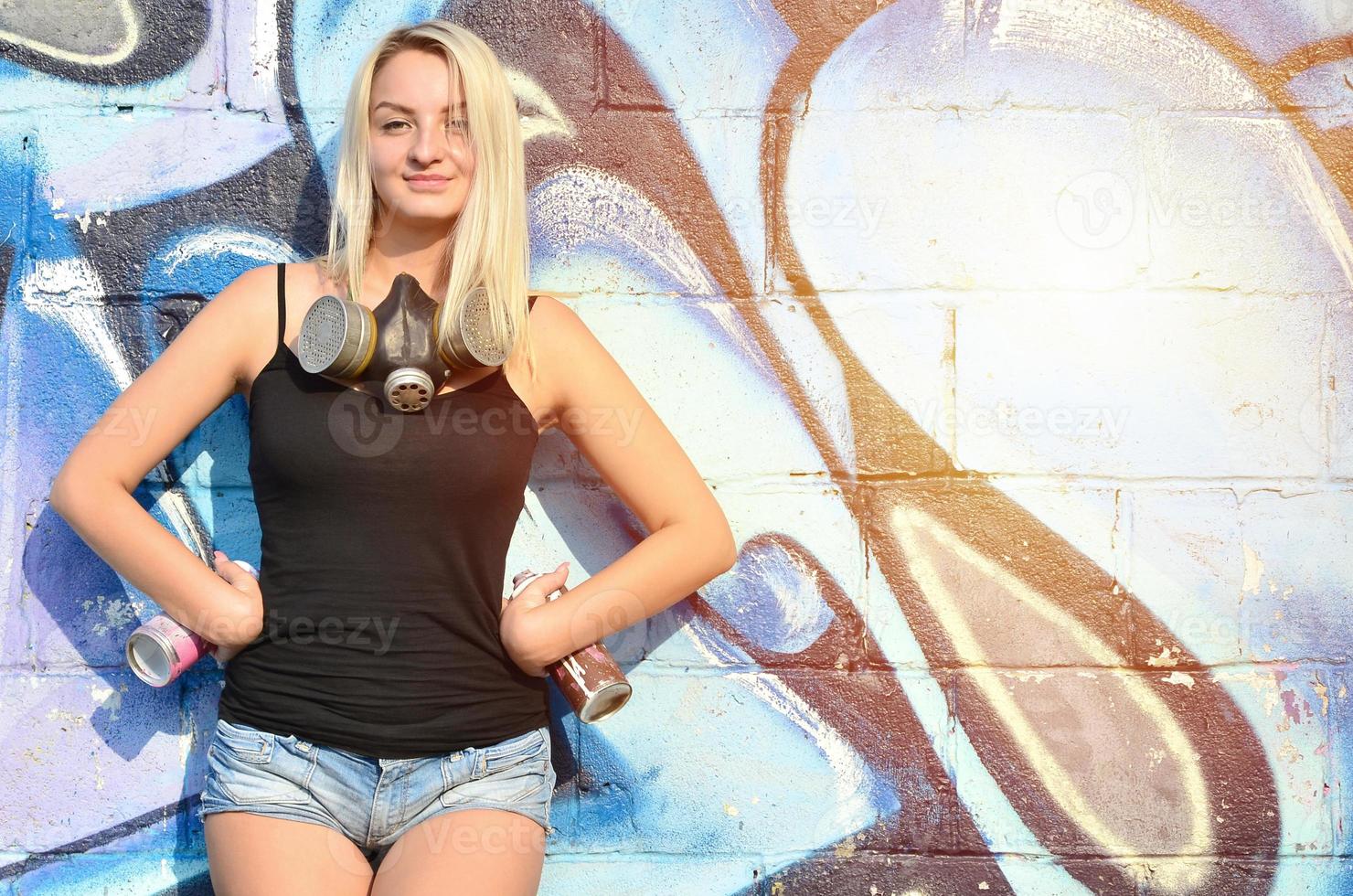 A smiling young and beautiful sexy girl graffiti artist with a paint spray and gas mask on her neck stands on the wall background with a graffiti pattern in blue and purple tones photo