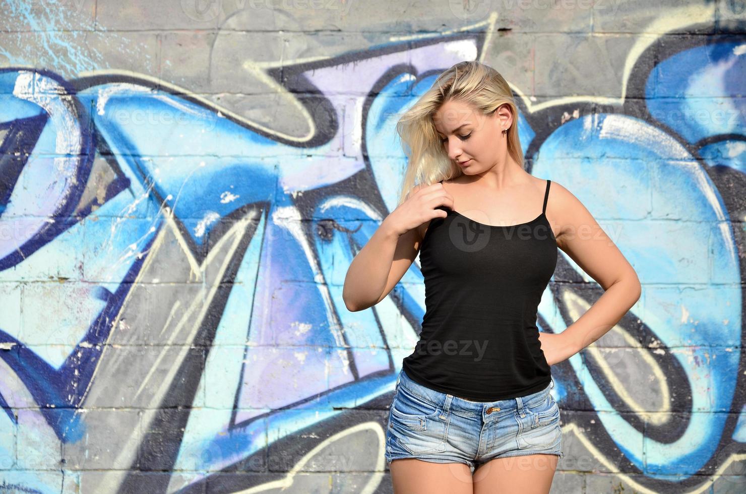 Chica rubia caucásica sexy en pantalones cortos de mezclilla y camiseta sin mangas negra posando contra la pared de graffiti durante el día al aire libre foto