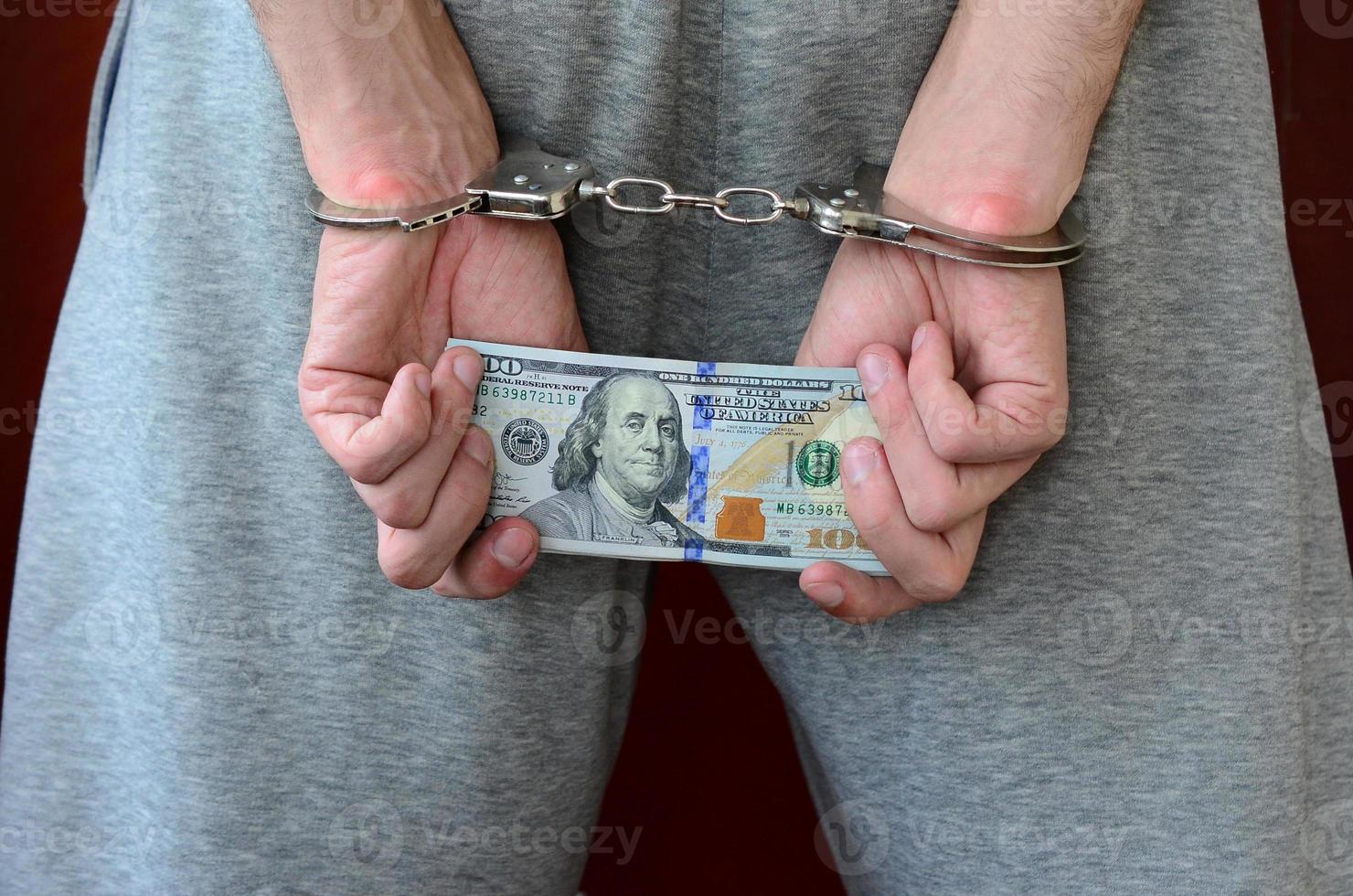 An arrested guy in gray pants with handcuffed hands holds a huge amount of dollar bills. Back view photo