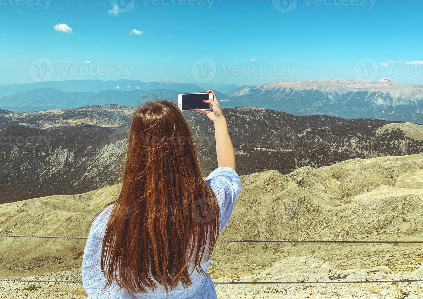 una chica con un vestido blanco y cabello largo y oscuro en la plataforma de observación se hace una selfie. foto tuya contra el telón de fondo de hermosas vistas de gran altura