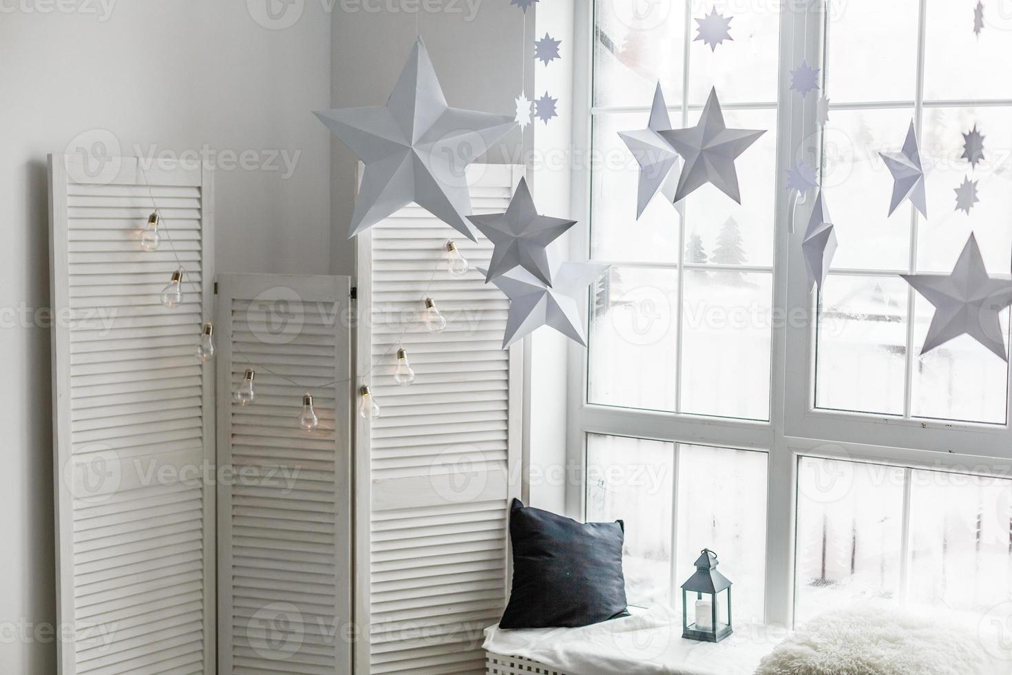 Large bright window. On the window-sill is a gray knitted plaid with gifts and sparkles with herlands. Paper Christmas stars are suspended on the background of the window. photo