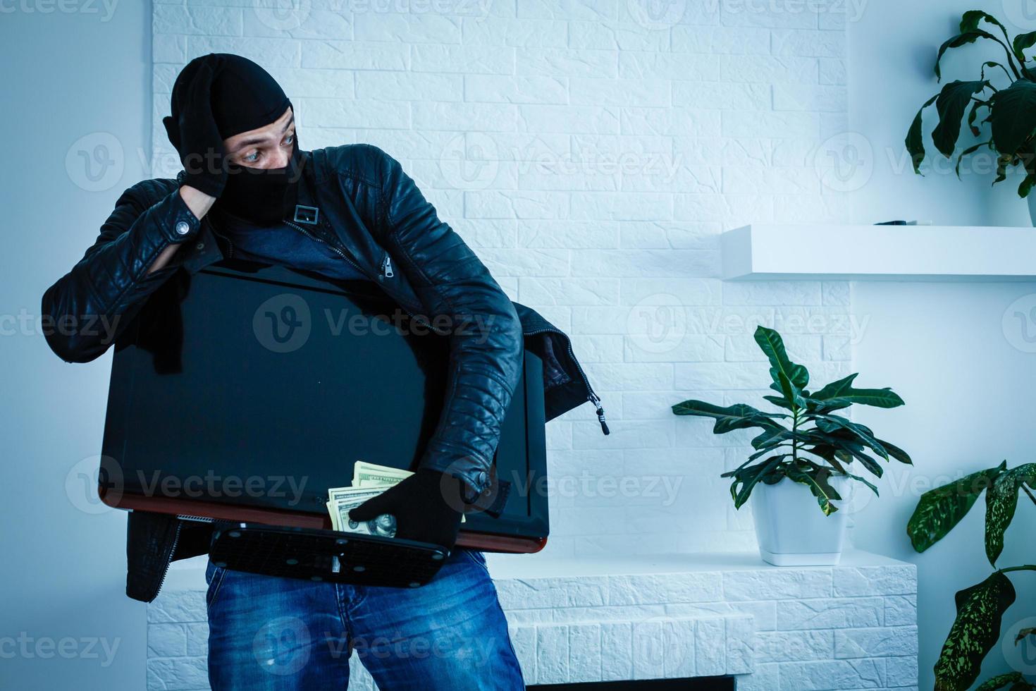 Robber man dressed in black hoodie stands with disguised face and holds a lot of money in his hands, stole a large amount, a thief man stole a TV photo