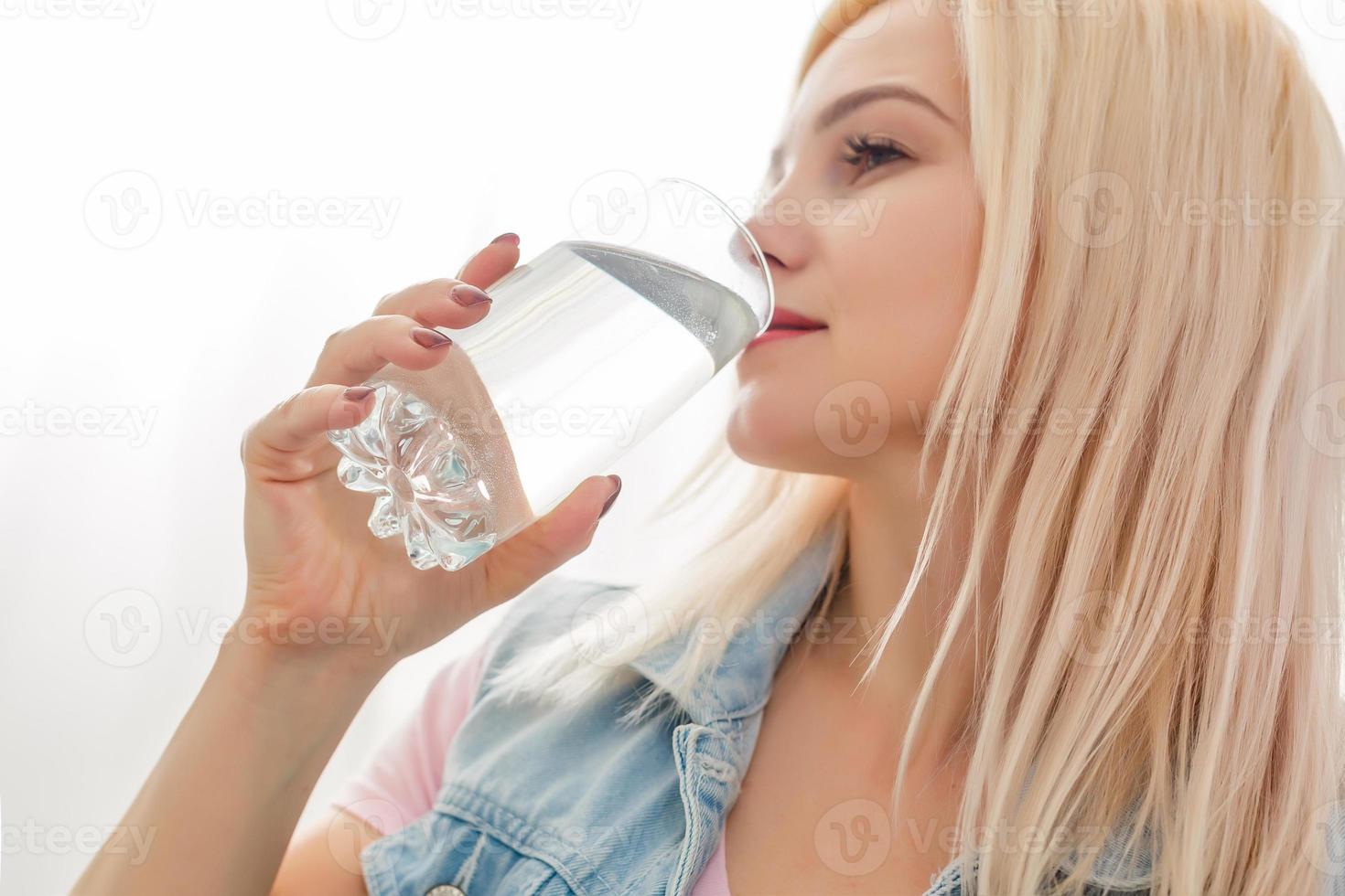 Happy beautiful young woman drinking water. Smiling caucasian female model holding transparent glass in her hand. Closeup. Focus on the arm photo