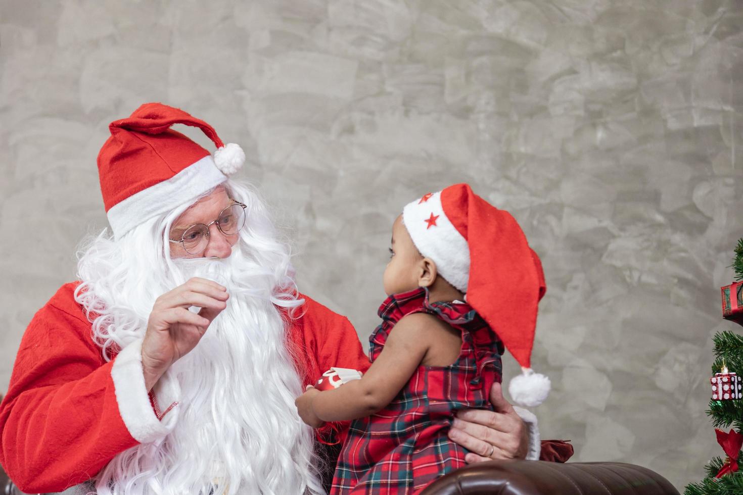 santa claus está levantando a una niña pequeña feliz y sentándose en su regazo con un árbol de navidad completamente decorado en la parte posterior para la celebración de la temporada foto