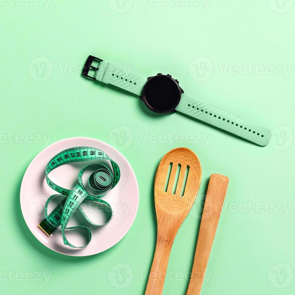 Diet sport concept, tape measure on a plate with wooden cultery and sport watch on a mint background. photo