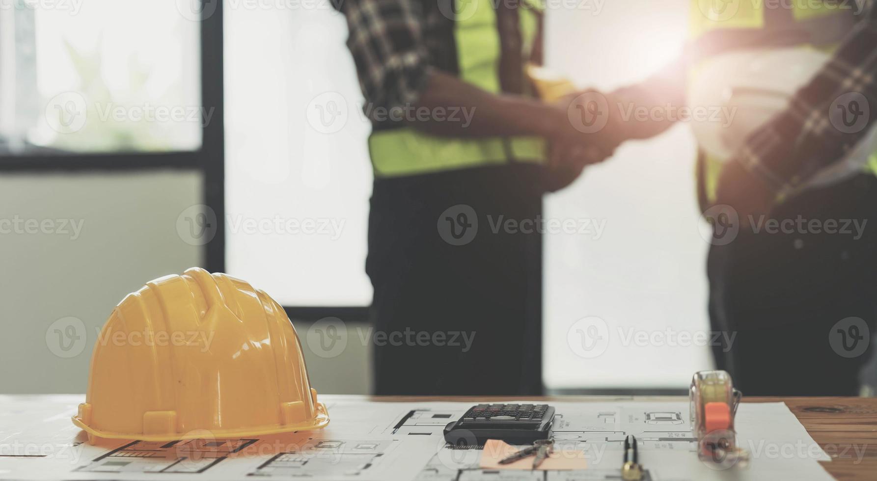 yellow safety helmet on workplace desk with construction worker team hands shaking greeting start up plan new project contract in office center at construction site, partnership and contractor concept photo