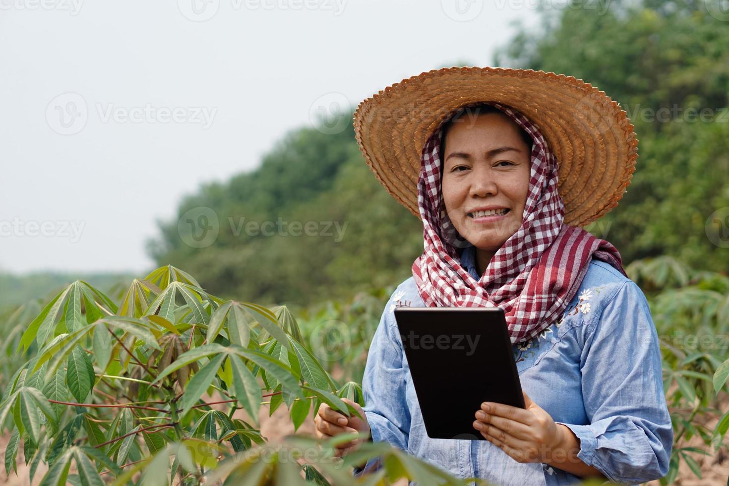 Portrait of Asian woman farmer, wears hat, Thai loincloth covered head, holds smart tablet at agriculture plantation. Concept, smart farmer, use technology internet to research and develop crops. photo