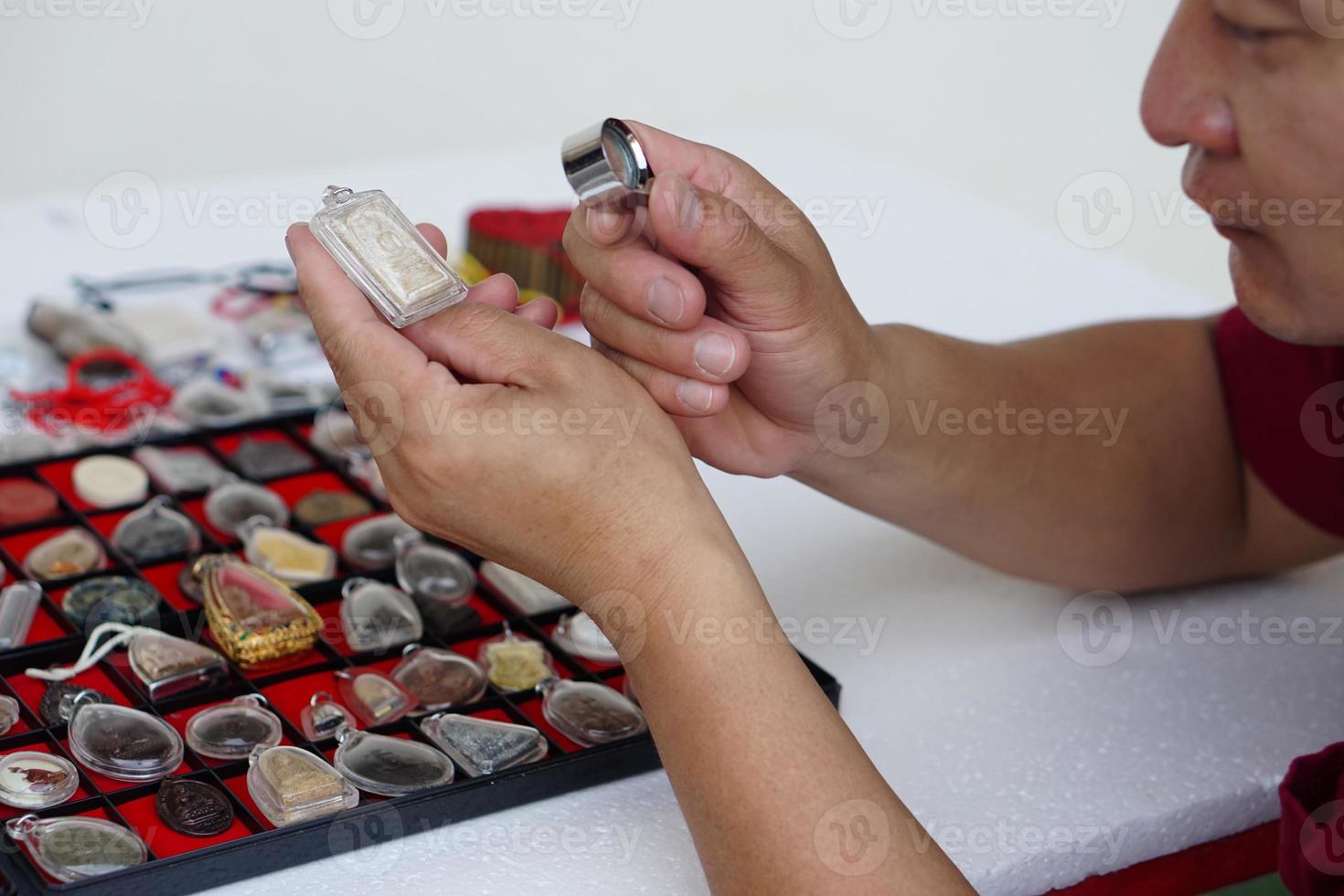 Closeup man's hands hold Buddha amulet and magnifying glass to check details and figure of object. Concept, faith, belief in holy and luck for Buddhists.  Colleting amulet as hobbies, exchange, trade. photo