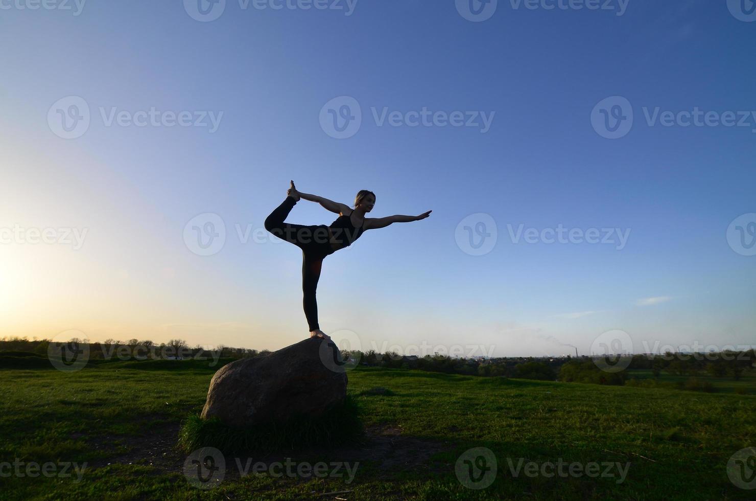 la silueta de una joven rubia con traje deportivo practica yoga en una pintoresca colina verde por la noche al atardecer. el concepto de ejercicio y estilos de vida saludables foto