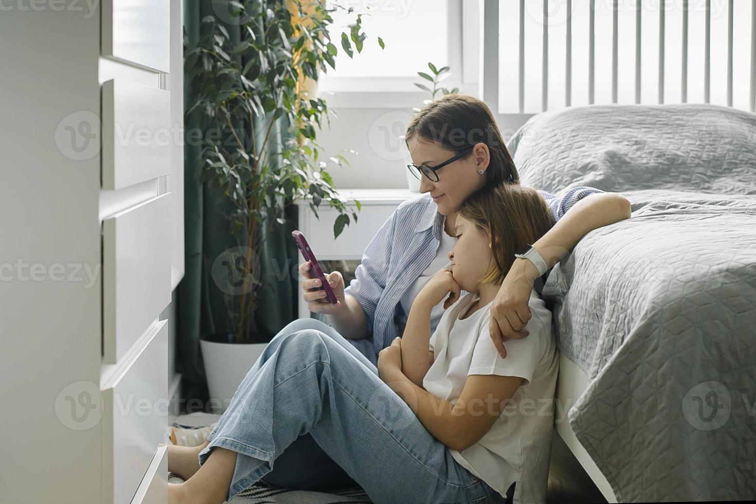 madre e hija usando un teléfono inteligente mientras están sentados en berdoom. mujer y niña preadolescente juntas foto