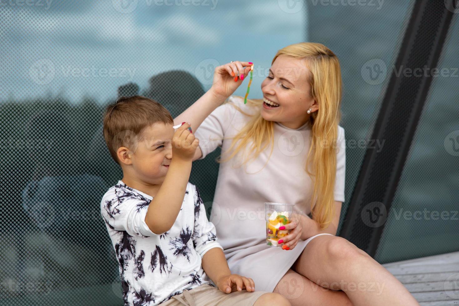 Happy blond woman and little boy sitting on terrace and eating sweets. Mother and son enjoy time together. Positive young mom playing, spending time with her cute child, laughing, having fun. Family. photo