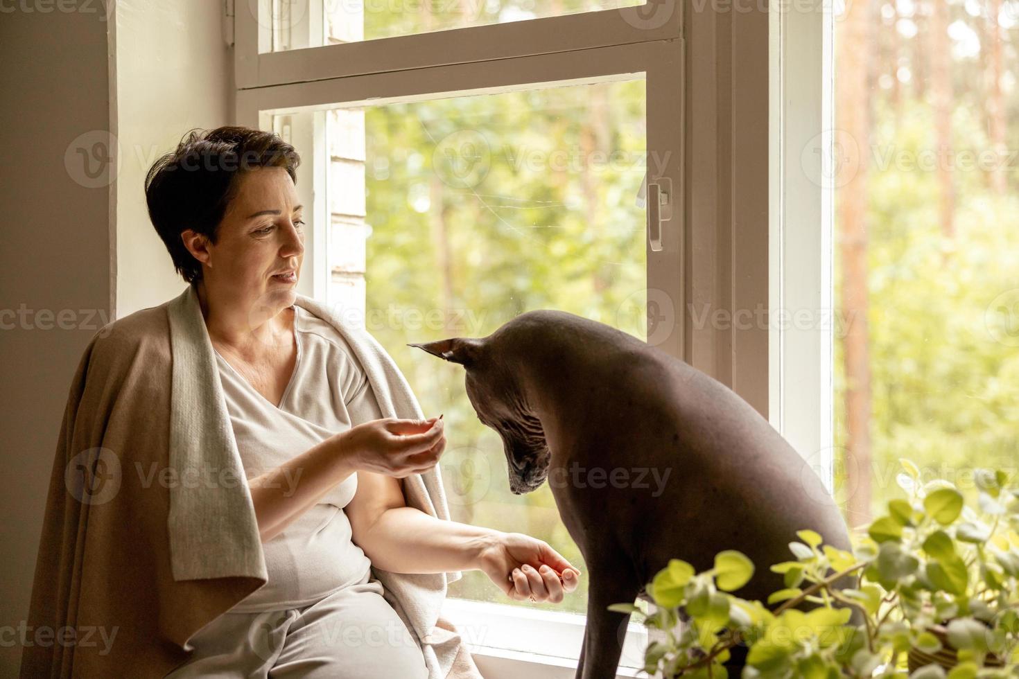 Middle age beautiful woman sitting on windowsill with her dog. 50-year-old woman spending time with her Mexican hairless dog at home. Xoloitzquintle, xolo breed. Dog as best friend, family member. photo