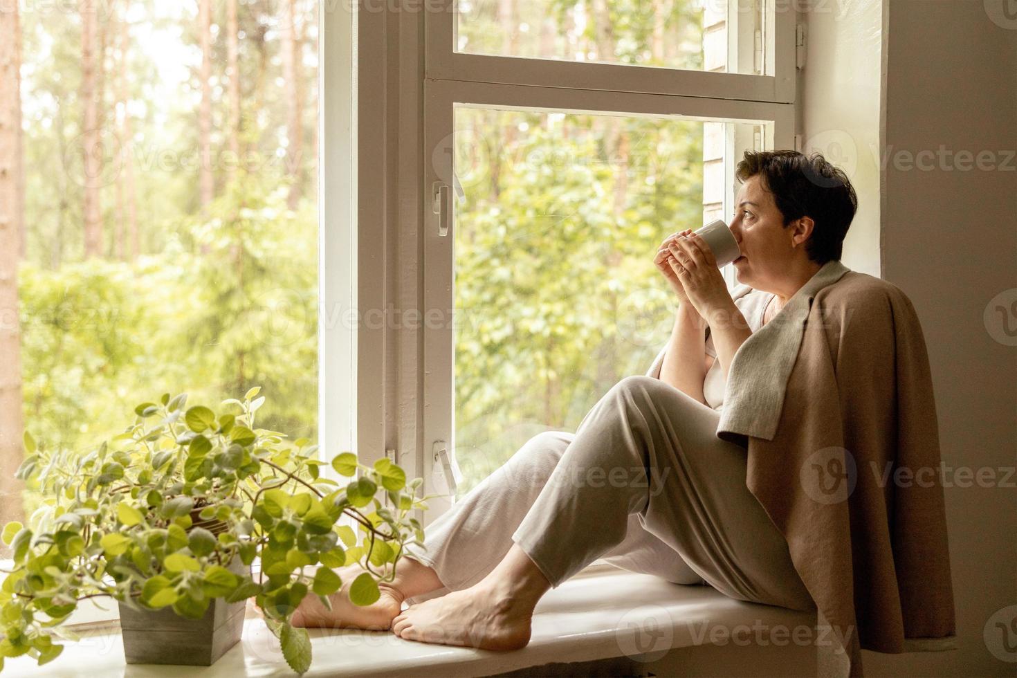 Middle age beautiful woman sitting on windowsill, drinking tea, dreaming. 50-year-old woman relaxing with cup of tea. Relax at home, looking away, thinking, stress free, peaceful mood, wellbeing alone photo