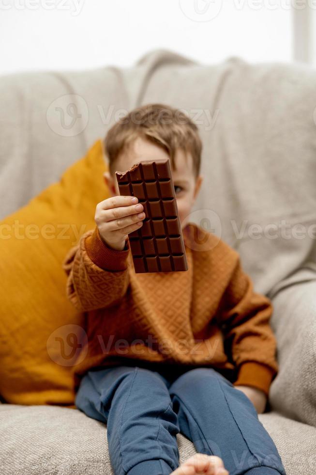 Little adorable boy sitting on the couch at home and eating chocolate bar. Child and sweets, sugar confectionery. Kid enjoy a delicious dessert. Preschool child with casual clothing. photo