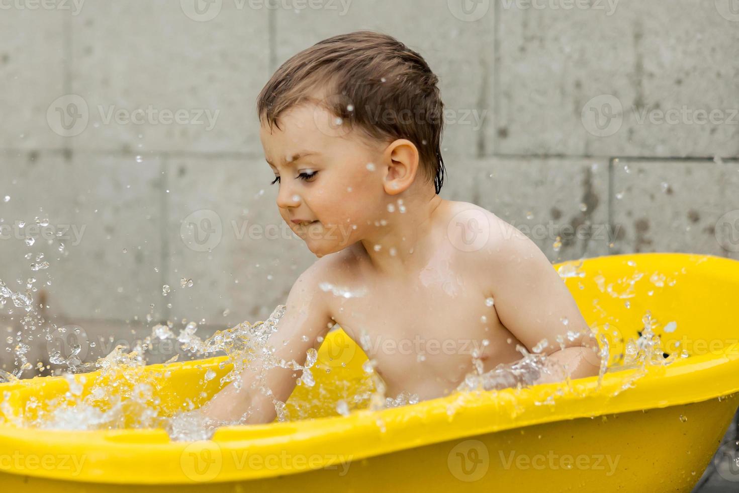 Cute little boy bathing in yellow tub outdoors. Happy child is splashing, playing with water and having fun. Summer season and recreation. Staying cool in the summer heat. Water fun in backyard. photo