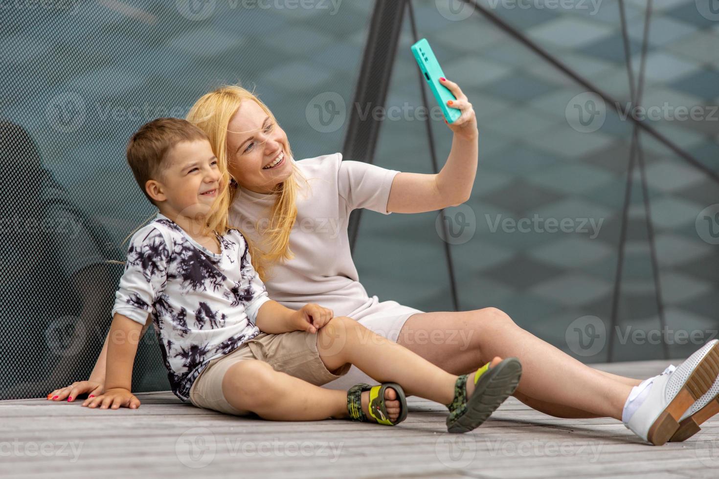 Happy blond woman and little boy sitting on terrace and making selfie on smartphone. Mother and son enjoy time together. Positive young mom spending time with her cute child. Having fun. Family. photo