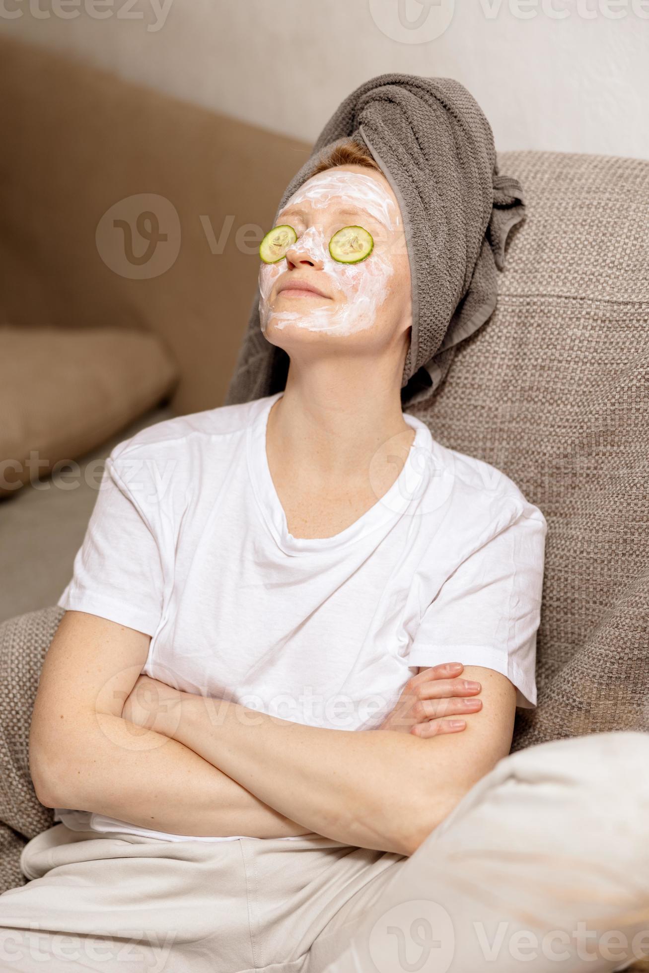 Young woman with casual clothes, towel on head and beauty mask on face sitting on couch and
