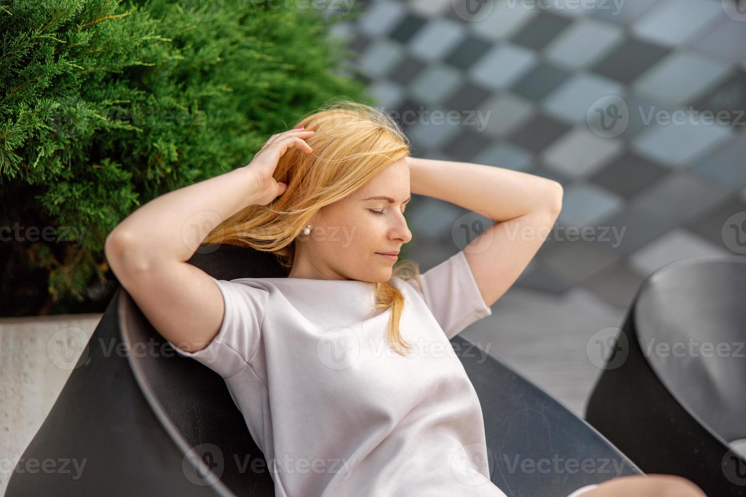 Young beautiful blond woman sitting outdoors, on the wooden terrace in the city and resting. Girl has break, spending time outside and relaxing. Time with yourself, dreaming, relaxation, mental health photo