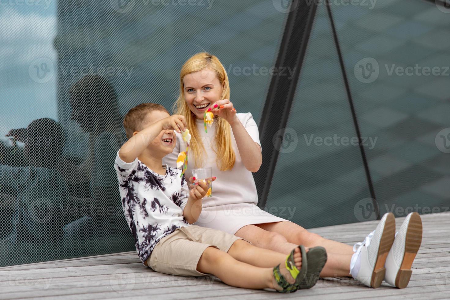 Happy blond woman and little boy sitting on terrace and eating sweets. Mother and son enjoy time together. Positive young mom playing, spending time with her cute child, laughing, having fun. Family. photo