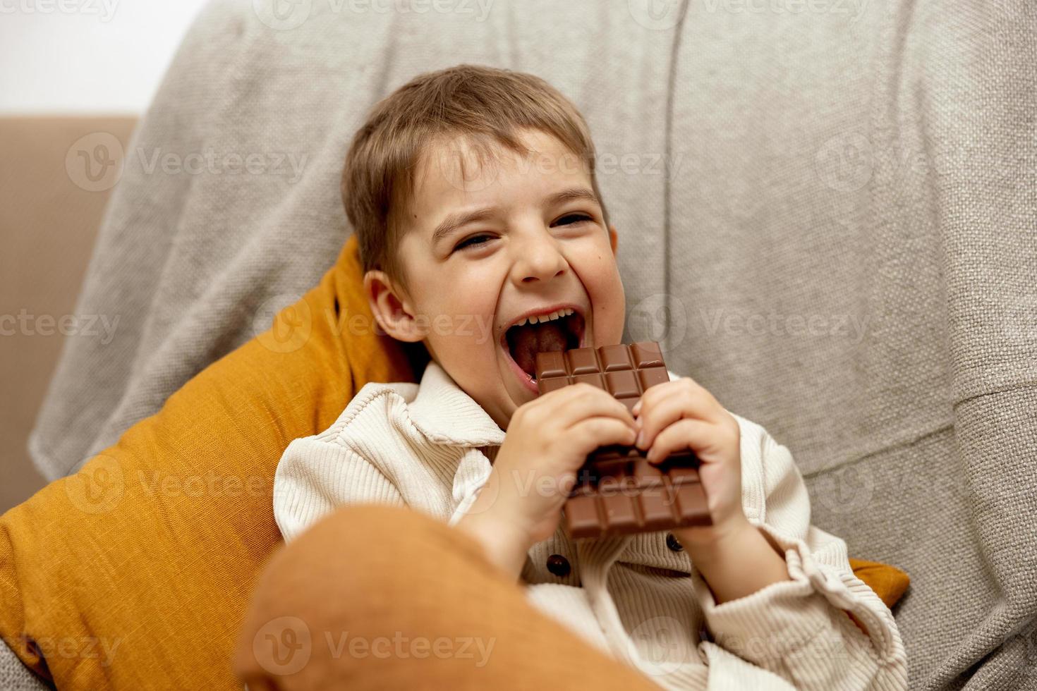 Little adorable boy sitting on the couch at home and eating chocolate bar. Child and sweets, sugar confectionery. Kid enjoy a delicious dessert. Preschool child with casual clothing. Positive emotion. photo