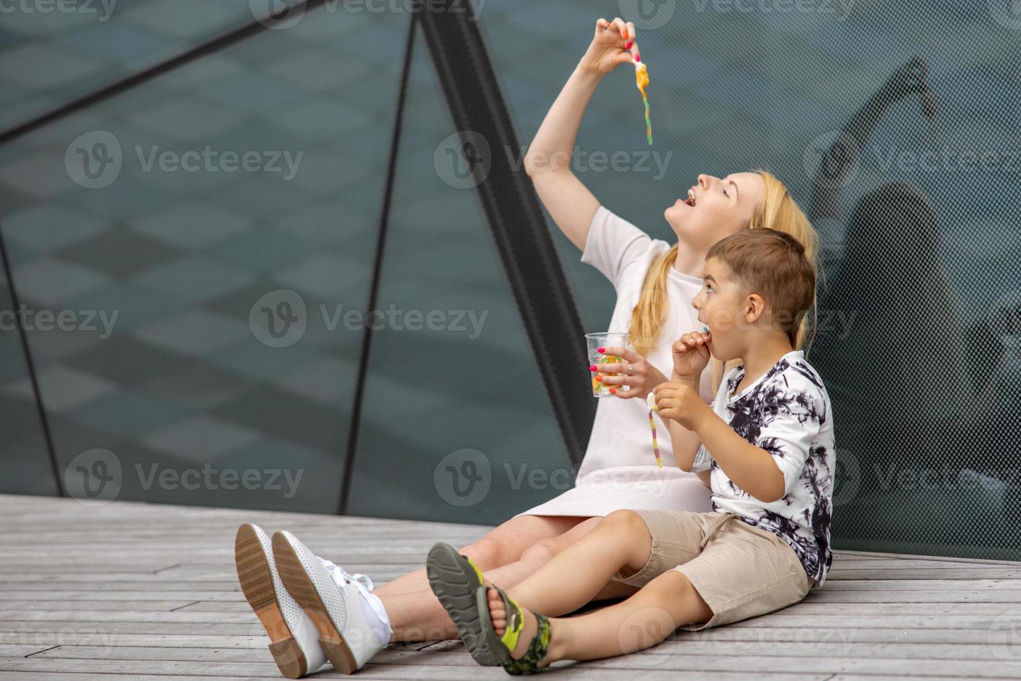 Happy blond woman and little boy sitting on terrace and eating sweets. Mother and son enjoy time together. Positive young mom playing, spending time with her cute child, laughing, having fun. Family. photo