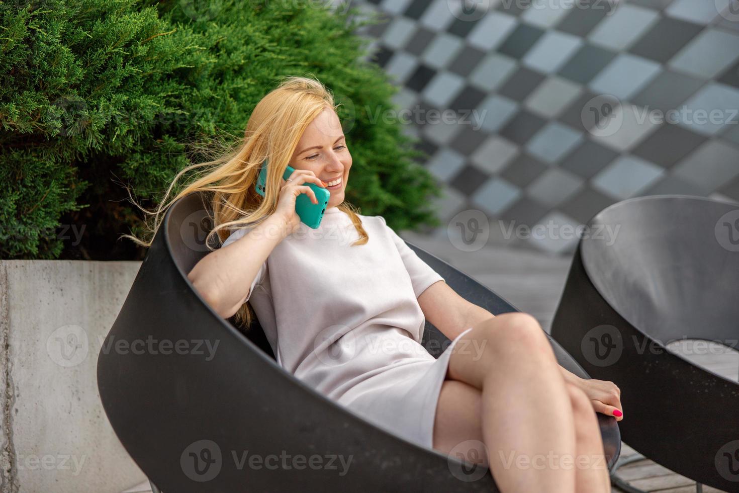 Young blond woman sittings outdoors in the city and making a call with her smartphone. Girl talking with friends with mobile phone. Leisure activity, communication. Conversation. Mobile technology. photo