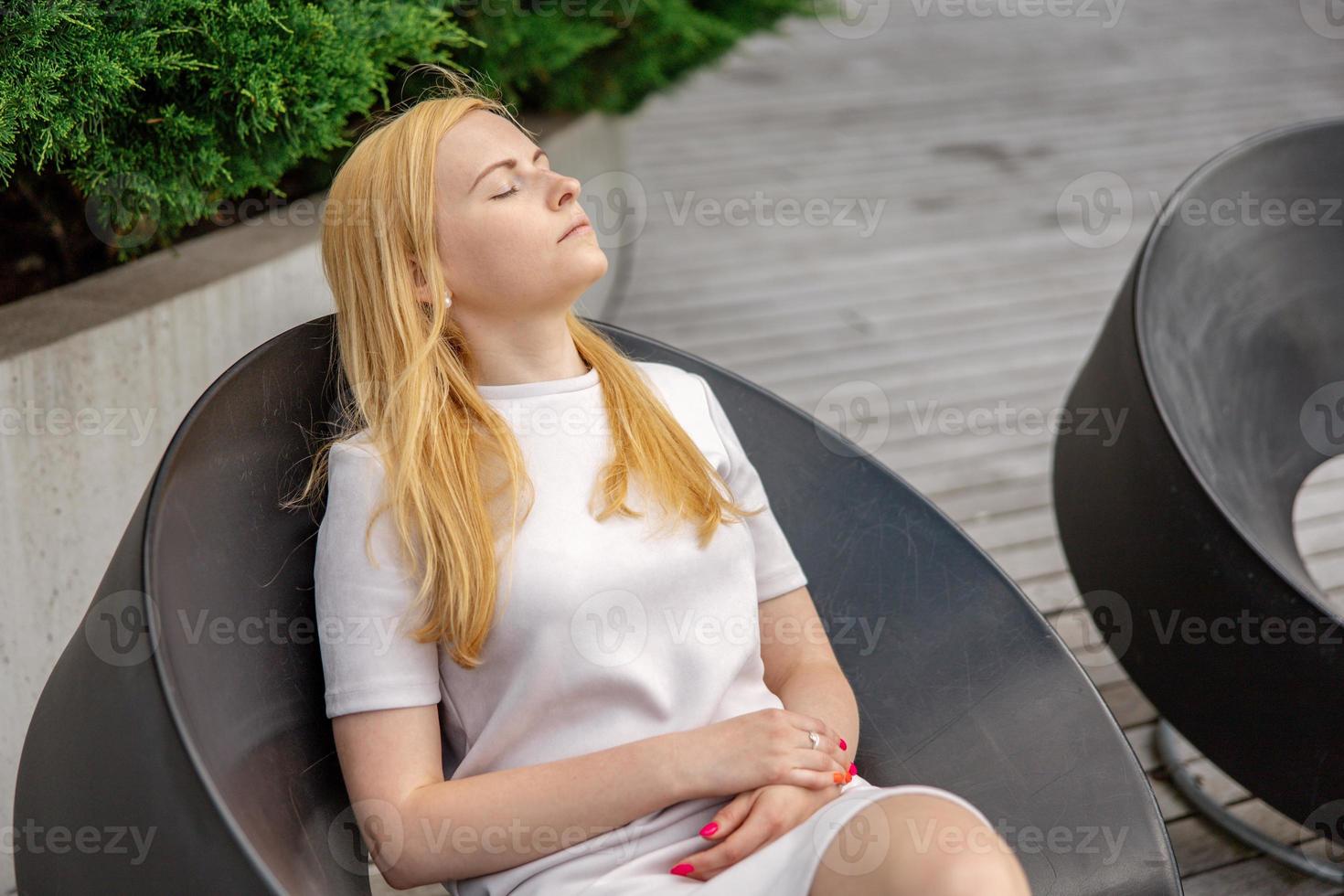 joven y bella mujer rubia sentada al aire libre, en la terraza de madera de la ciudad y descansando. la chica tiene descanso, pasa tiempo al aire libre y se relaja. tiempo contigo mismo, soñar, relajación, salud mental foto