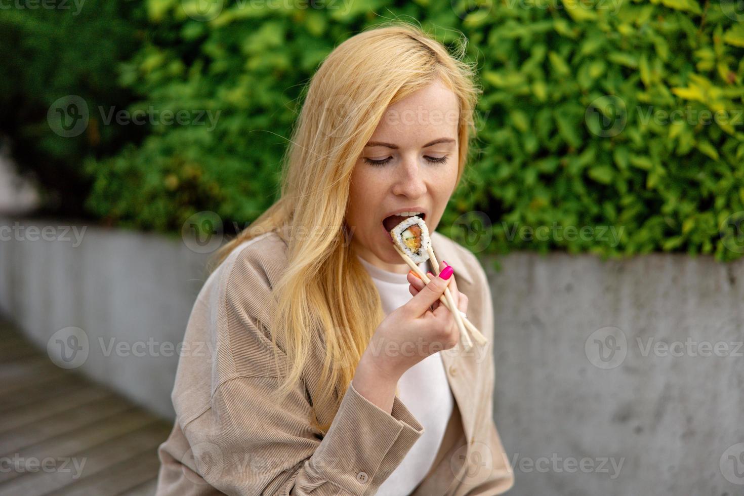 Young beautiful blond woman eating sushi outdoors, on the wooden terrace, by modern building in the city. Tasty food to go. Girl has lunch break, spending time outside and eating Asian food. City life photo