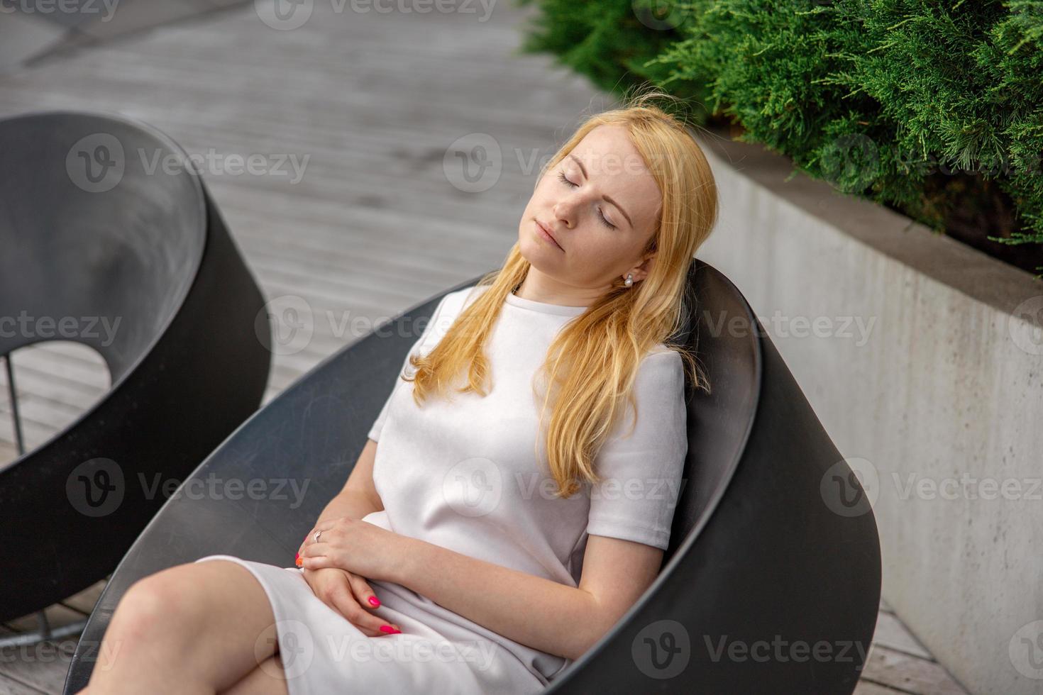 Young beautiful blond woman sitting outdoors, on the wooden terrace in the city and resting. Girl has break, spending time outside and relaxing. Time with yourself, dreaming, relaxation, mental health photo