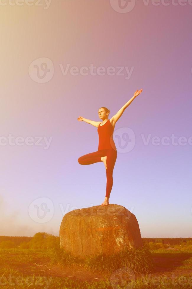 la silueta de una joven rubia con traje deportivo practica yoga en una pintoresca colina verde por la noche al atardecer. el concepto de ejercicio y estilos de vida saludables foto